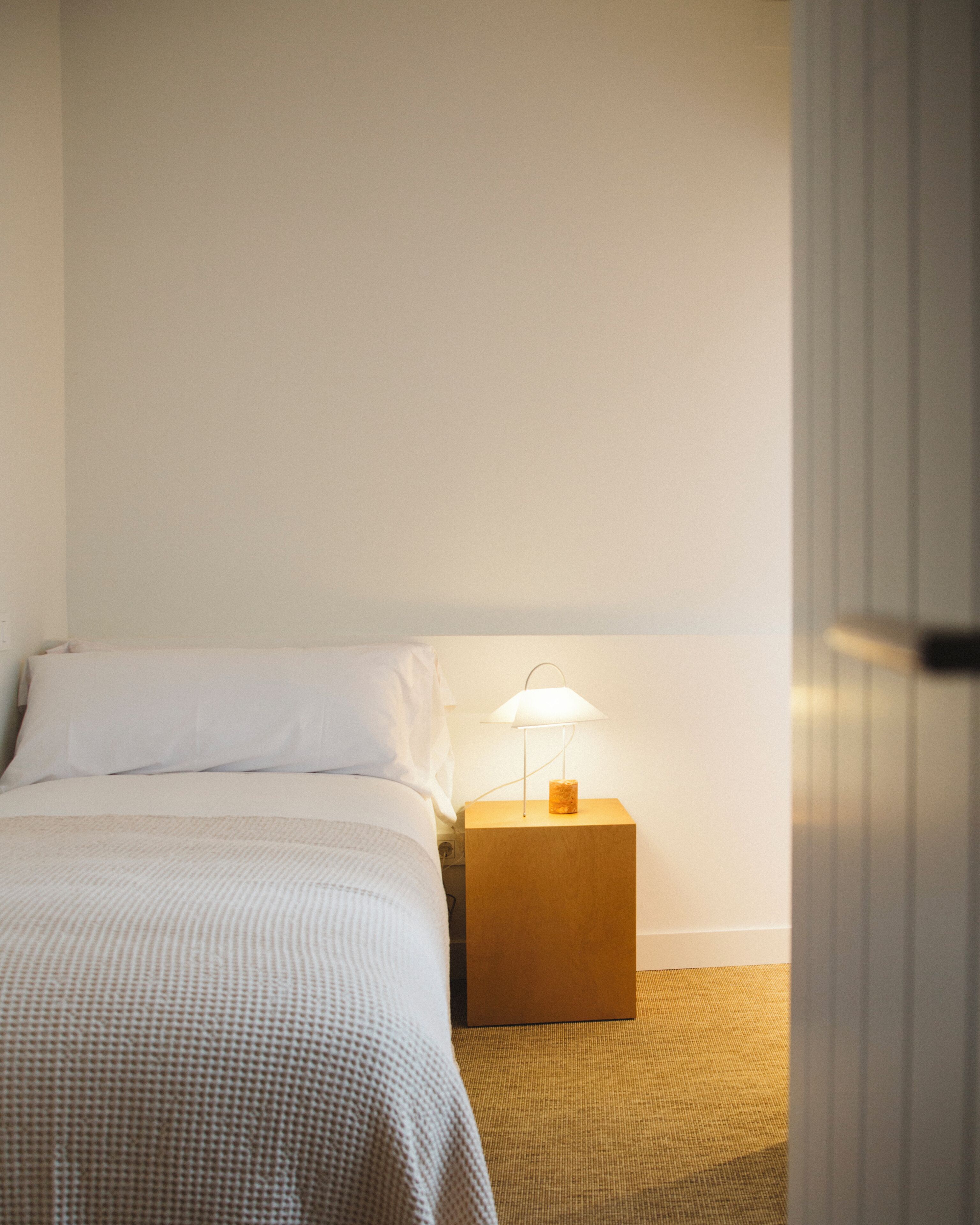 A hotel room with a bed, a nightstand with a lamp, and a door.