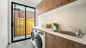 A laundry area with a washing machine, a sink, wooden cabinets, and a sliding glass door leading to a fenced outdoor area. - Park-fronted Luxury Grand Family House with Gym, Heated Pool/Spa, Tennis Court. (Mambourin)