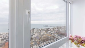 Janela com vista para uma cidade costeira, um navio ao longe e um vaso com flores no parapeito.