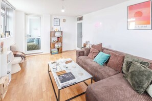 Ein Wohnzimmer mit einem braunen Sofa, einem weißen Couchtisch mit Büchern, einem Holzregal und einem gerahmten Bild an der Wand.