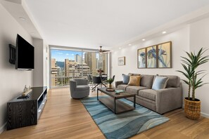 A modern living room with a sofa, coffee table, TV, and a large window with a city view. - 14th Floor Waikiki City & Mountain Views! Full Kitchen! Walk to Beach! (Honolulu)