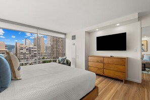 A bedroom with a large bed, a wooden dresser, a flat-screen TV, and a city view. - 14th Floor Waikiki City & Mountain Views! Full Kitchen! Walk to Beach! (Honolulu)