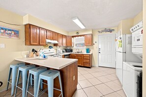 A kitchen with wooden cabinets, a central island with stools, a microwave, stove, and white appliances. - 2BR with loft, sleeps 8, lagoon front, pool & private beach access (Gulf Shores)