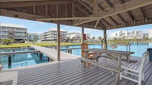 A wooden deck with a pool, picnic tables, and a covered area overlooking a marina with boats and buildings. - 2BR with loft, sleeps 8, lagoon front, pool & private beach access (Gulf Shores)