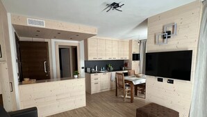 A modern kitchen with wooden cabinets, a dining area with a table and chairs, and a large TV mounted on the wall. - Bormio Apartments al pradais  giardino est  (Bormio)