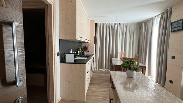A modern kitchen with wooden cabinets, a countertop, and a dining area with a table and chairs. - Bormio Apartments al pradais  giardino est  (Bormio)