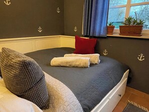 Ein Bett mit dunkler Decke, einem roten Kissen und zwei Handtüchern. Auf einem Fensterbrett stehen Blumentöpfe.