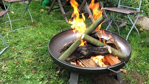 Ulkoterassi, jossa on nuotiopaikka, pöytä, jossa on lautasia, sekä kahvipannu ja raidallinen aurinkovarjo.