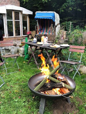 Ein Garten mit Feuerstelle, einem Tisch gedeckt mit Speisen und einem Streifen-Sonnenschirm.
