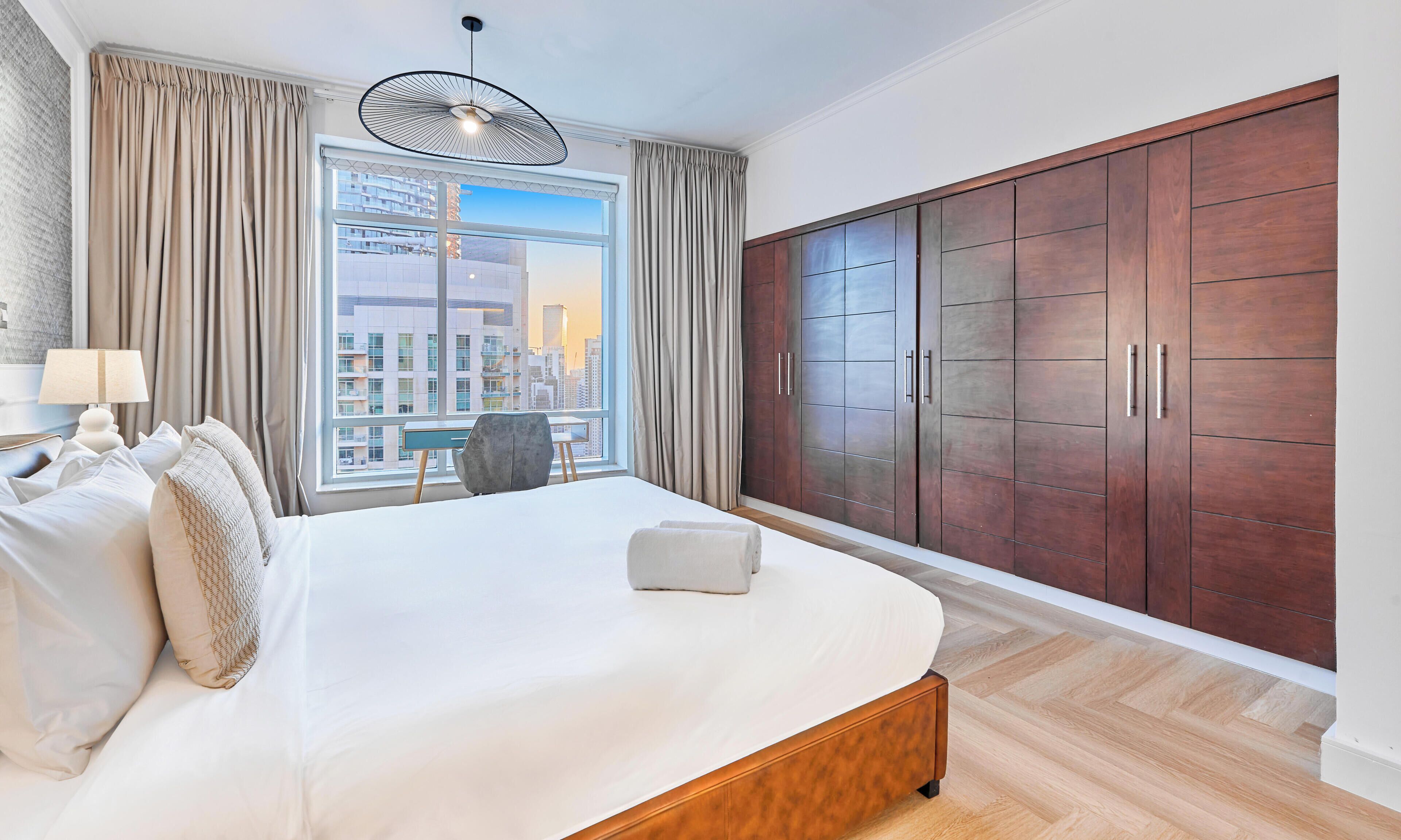 A hotel room with a large bed, a desk, a chair, a wardrobe, and a view of the cityscape.