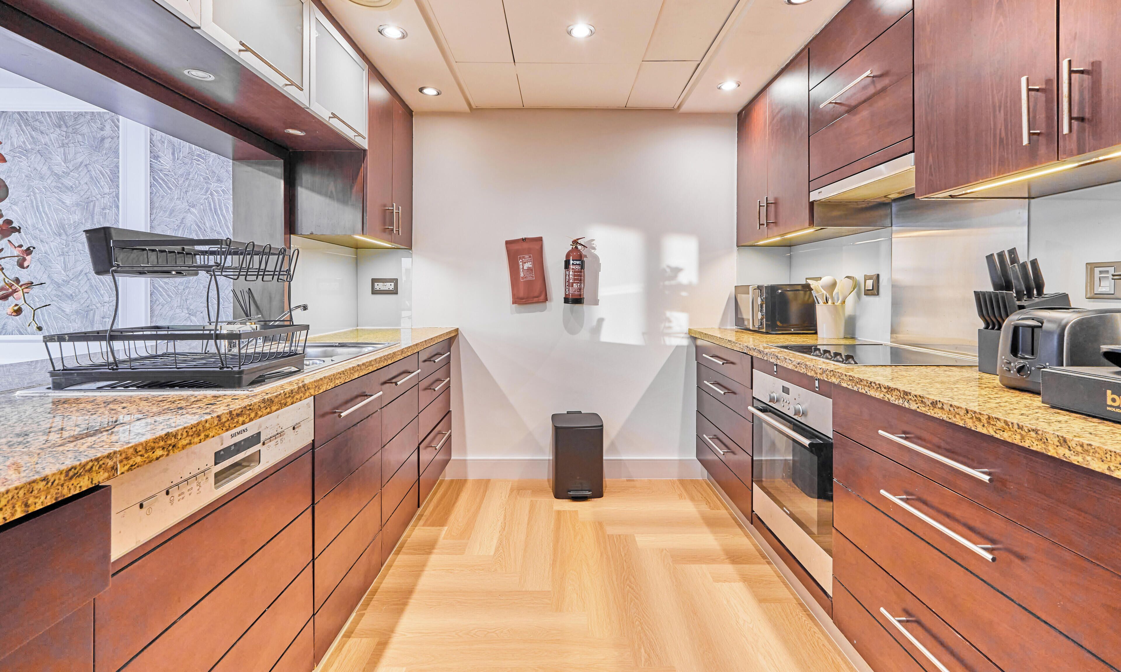Una cocina moderna con armarios de madera, encimeras de granito, lavavajillas Siemens, un cubo de basura negro y un extintor en la pared.