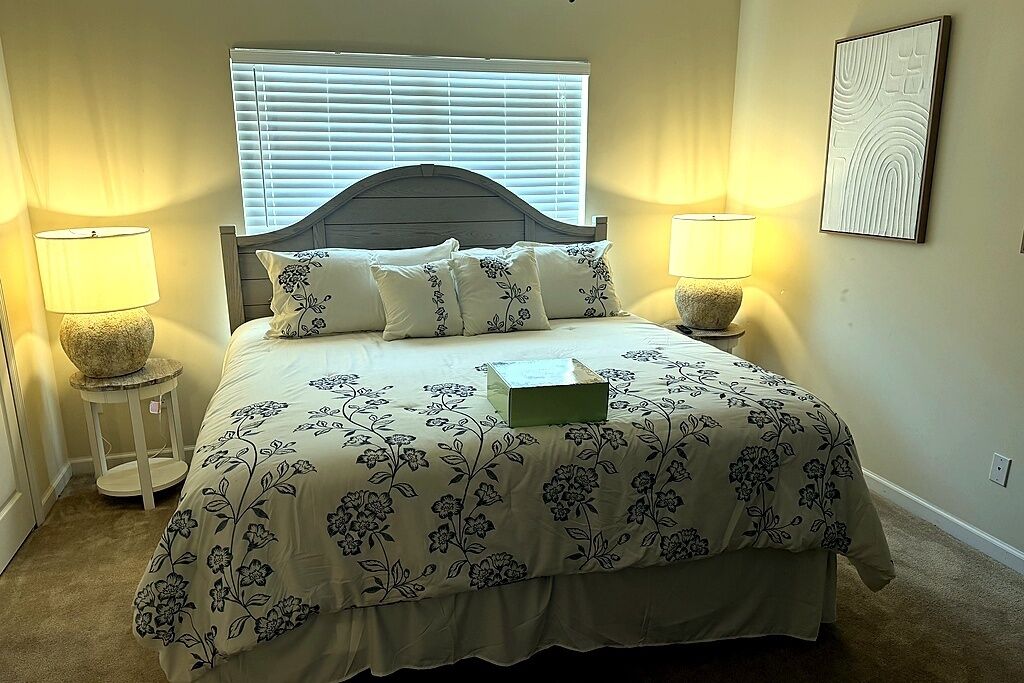 A bedroom with a bed, two bedside lamps, a window with blinds, and a wall-mounted artwork.