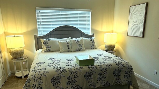 Un dormitorio con cama, dos lámparas de noche, una ventana con persianas y una obra de arte colgada en la pared.