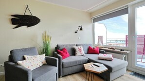 Una sala de estar moderna con sofás grises, una mesa de centro de madera, una decoración de peces en la pared y una ventana con vistas.