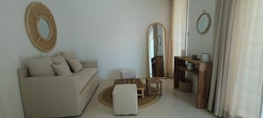 A living room with a sofa, a coffee table, a small table, a mirror, a wooden shelf, and a window with curtains.