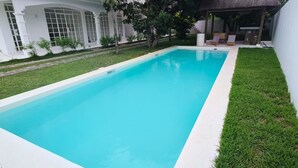 A rectangular swimming pool with a white edge, surrounded by a well-maintained lawn and a white building with large windows. - La Maison Coloniale II (Grand Baie)