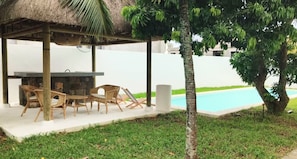 Ein überdachter Außenbereich mit Stühlen, Tisch, Pool und einer Palme.