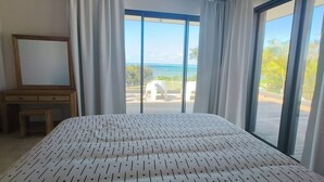 Un dormitorio con una cama grande, un aparador de madera, un espejo y vista al mar a través de ventanales.