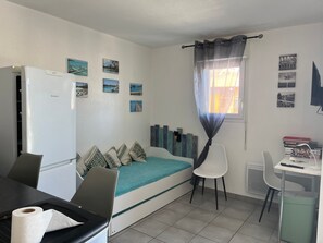 Ein kleines Zimmer mit Bett, Schreibtisch, Stuhl, Kühlschrank und einem fensterbelagten Fenster.
