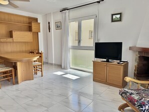A room with a wooden dining table, chairs, a television on a cabinet, a fireplace, and a large window with curtains. - Spacious apartment, sea view - 4 guests (Roses)