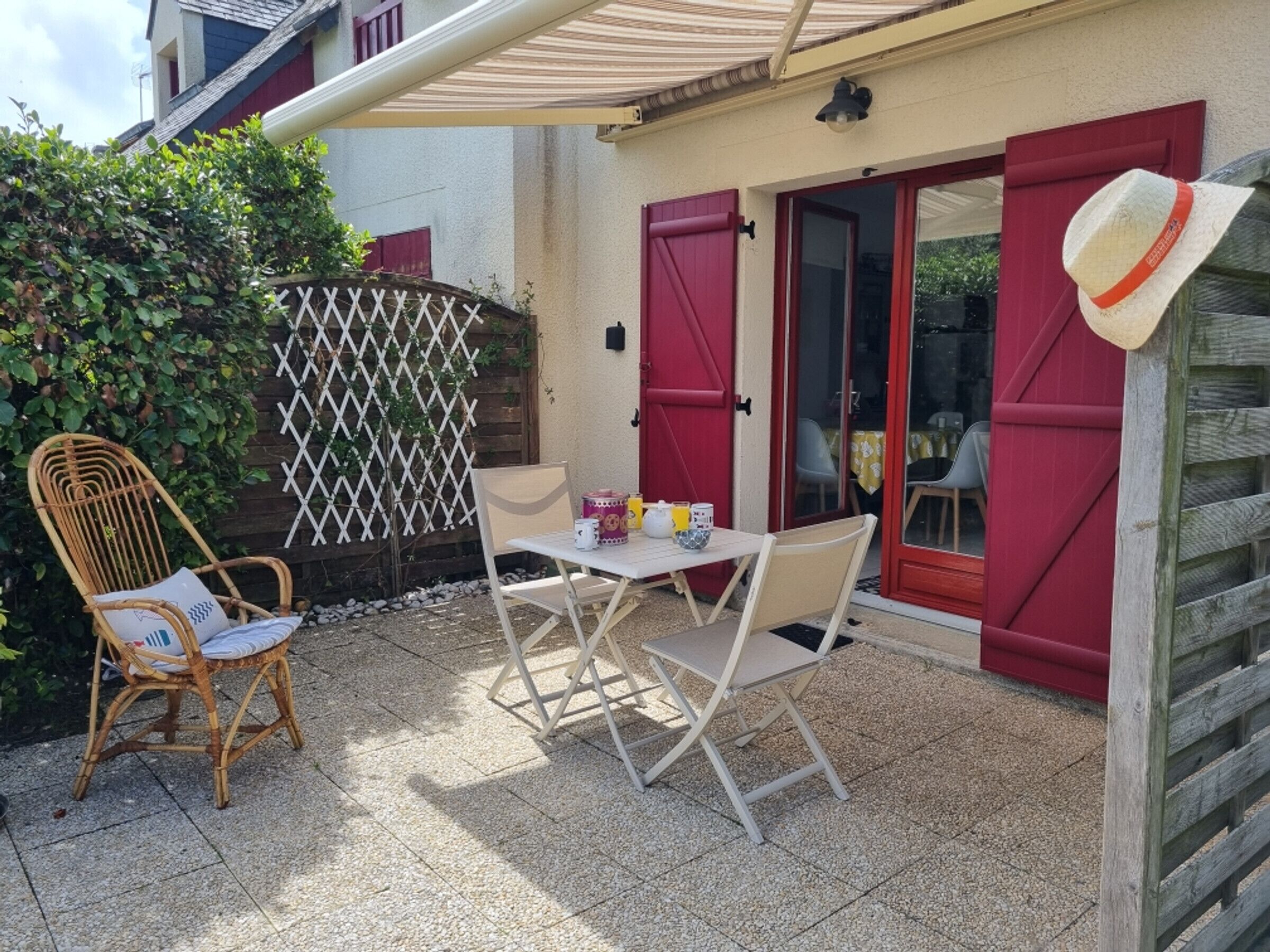 A patio with a table, two chairs, a wicker chair, a red door, and a white fence.