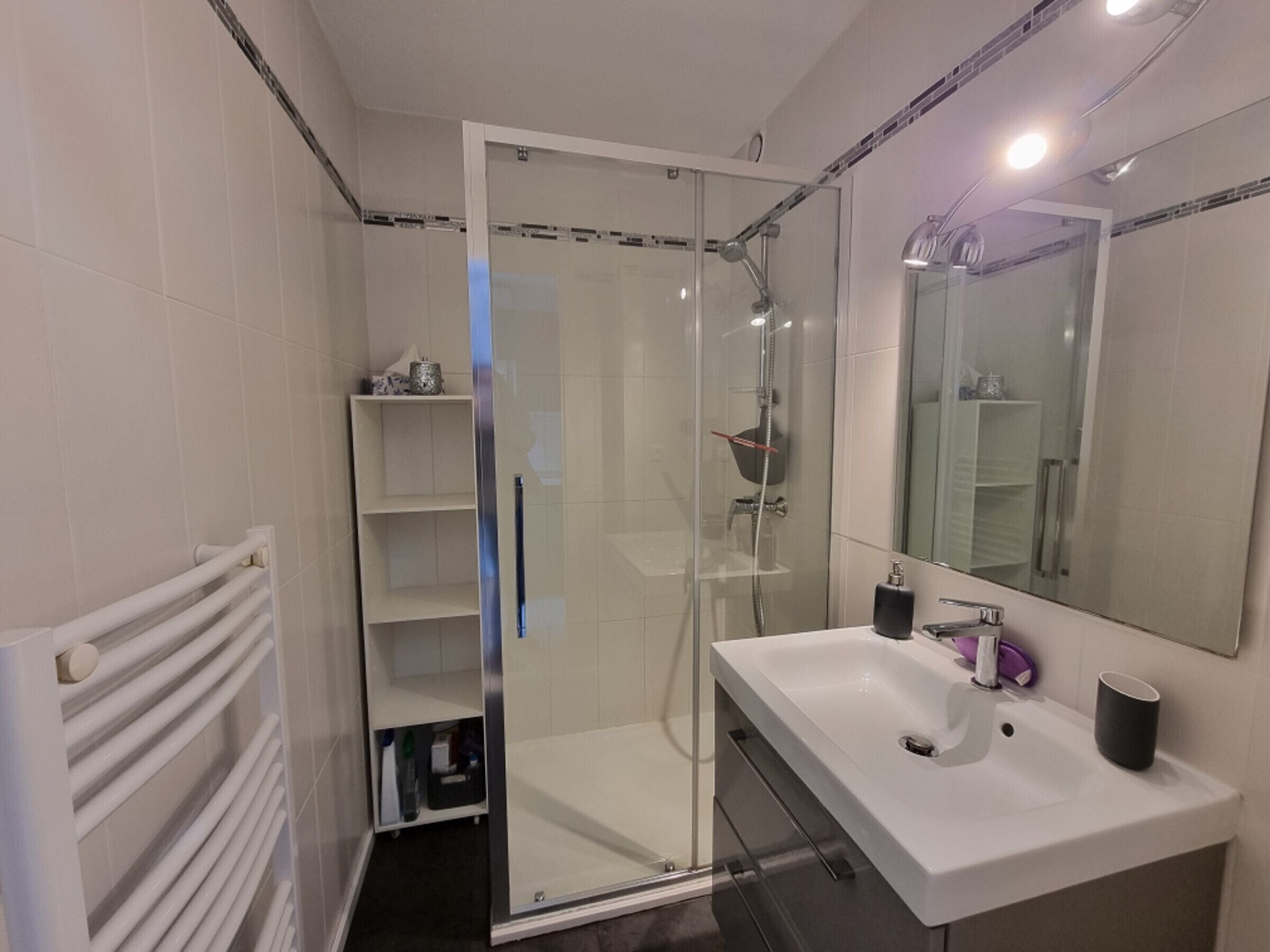 Een moderne badkamer met een douche met glazen wand, een wit wastafeltje, een spiegel en een handdoekrek.