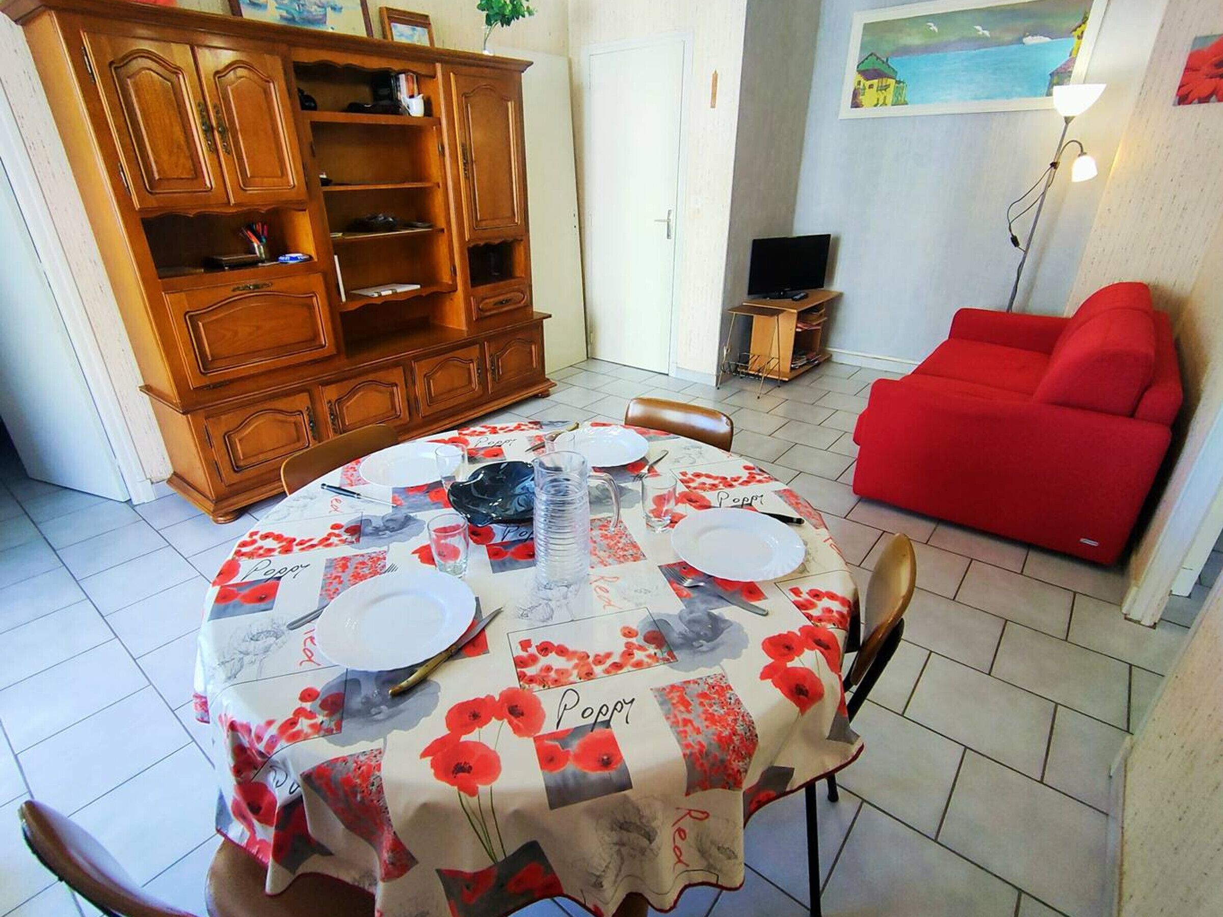 A dining area with a round table set for two, a wooden cabinet, a red sofa, and a television in the background.
