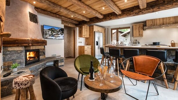 Ein rustikales Interieur mit Kamin aus Stein, Holzbalkendecke, einer Essecke mit Barhocker und einer Küche im Hintergrund.