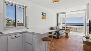 Eine moderne Küche mit Geschirrspüler, Barhocker, grauem Sofa und Esstisch mit Stühlen. Ein großes Fenster bietet Meerblick, und an der Wand hängt ein Gemälde.