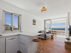 Eine moderne Küche mit Geschirrspüler, Barhocker, grauem Sofa und Esstisch mit Stühlen. Ein großes Fenster bietet Meerblick, und an der Wand hängt ein Gemälde.