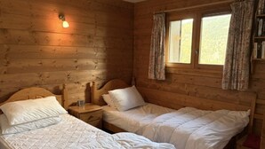 Puinen mökkimainen huone, jossa on kaksi sänkyä, yöpöytä, ikkuna verhoilla ja seinään kiinnitetty valaisin.