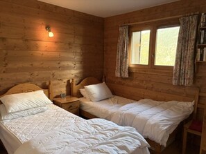A wooden cabin room with two beds, a nightstand, a window with curtains, and a wall-mounted light. - Apartment with slope view - 6 guests (Les Carroz d'Arâches)