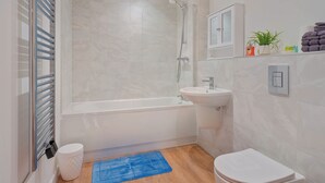 Moderni kylpyhuone, jossa on kylpyamme, pesuallas, wc-istuin, sininen matto ja kaappi, jossa on hygieniatuotteita.