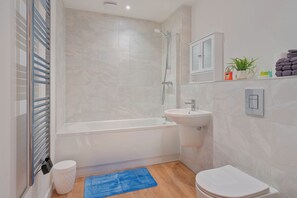 A modern bathroom with a bathtub, a sink, a toilet, a blue rug, and a cabinet with toiletries. - City Point Aberdeen by Guestz (Aberdeen)