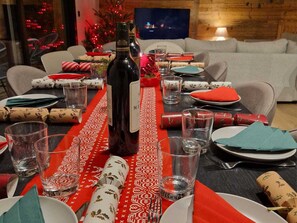 Ein Esstisch ist gedeckt mit Tellern, Gläsern und einer Flasche, umgeben von Stühlen und einem Weihnachtsbaum im Hintergrund.