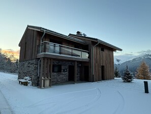 A two-story wooden cabin with a stone foundation, a balcony, and a view of snow-covered mountains. - Chalet with mountain view - 12 guests (La Plagne Montalbert)