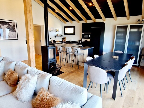 A modern living room with a sofa, dining table, and chairs, a kitchen area with a stove, and a large window. - Chalet with mountain view - 12 guests (La Plagne Montalbert)