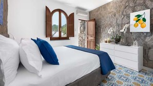 Una camera da letto con un letto, biancheria azzurra e bianca, un muro in pietra, una porta in legno, un comò bianco e un quadro con limoni.
