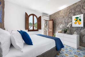 Un dormitorio con cama, ropa de cama blanca y azul, pared de piedra, puerta de madera, cómoda blanca y un cuadro de limones.