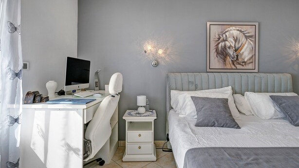 Quarto com cama, mesa de cabeceira, escrivaninha com computador e luminária na parede.