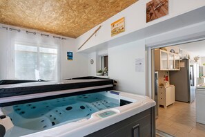 A spa room with a hot tub, wooden ceiling, and a view into the kitchen area.
