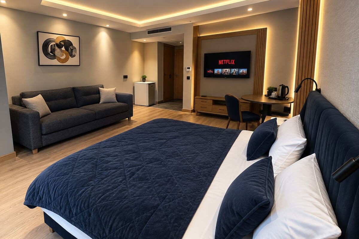 A modern hotel room with a large bed, a sofa, a desk with a computer, and a TV displaying Netflix.