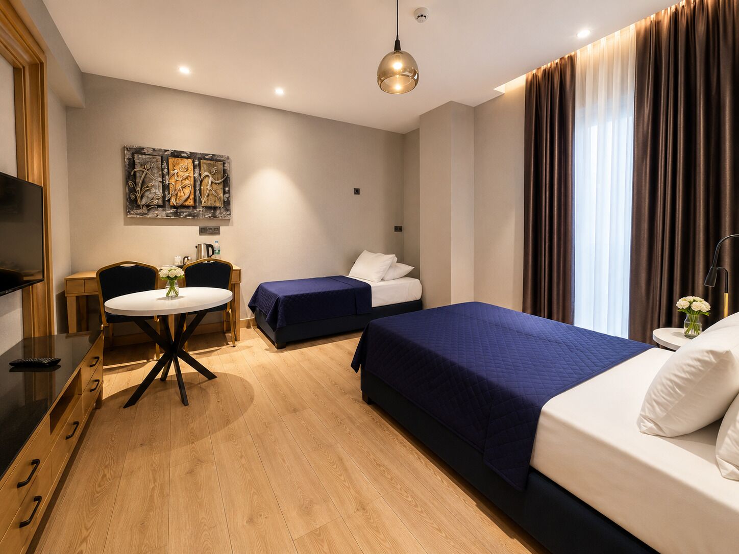 A hotel room with a bed, a desk with a chair, a small table with a vase of flowers, and a painting on the wall.
