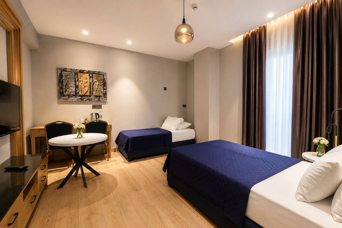 A hotel room with a bed, a desk with a chair, a small table with a vase of flowers, and a painting on the wall.