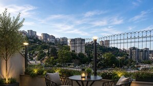 Una terraza en la azotea con una mesa redonda y dos sillas, rodeada de plantas en macetas y vistas a un paisaje urbano.