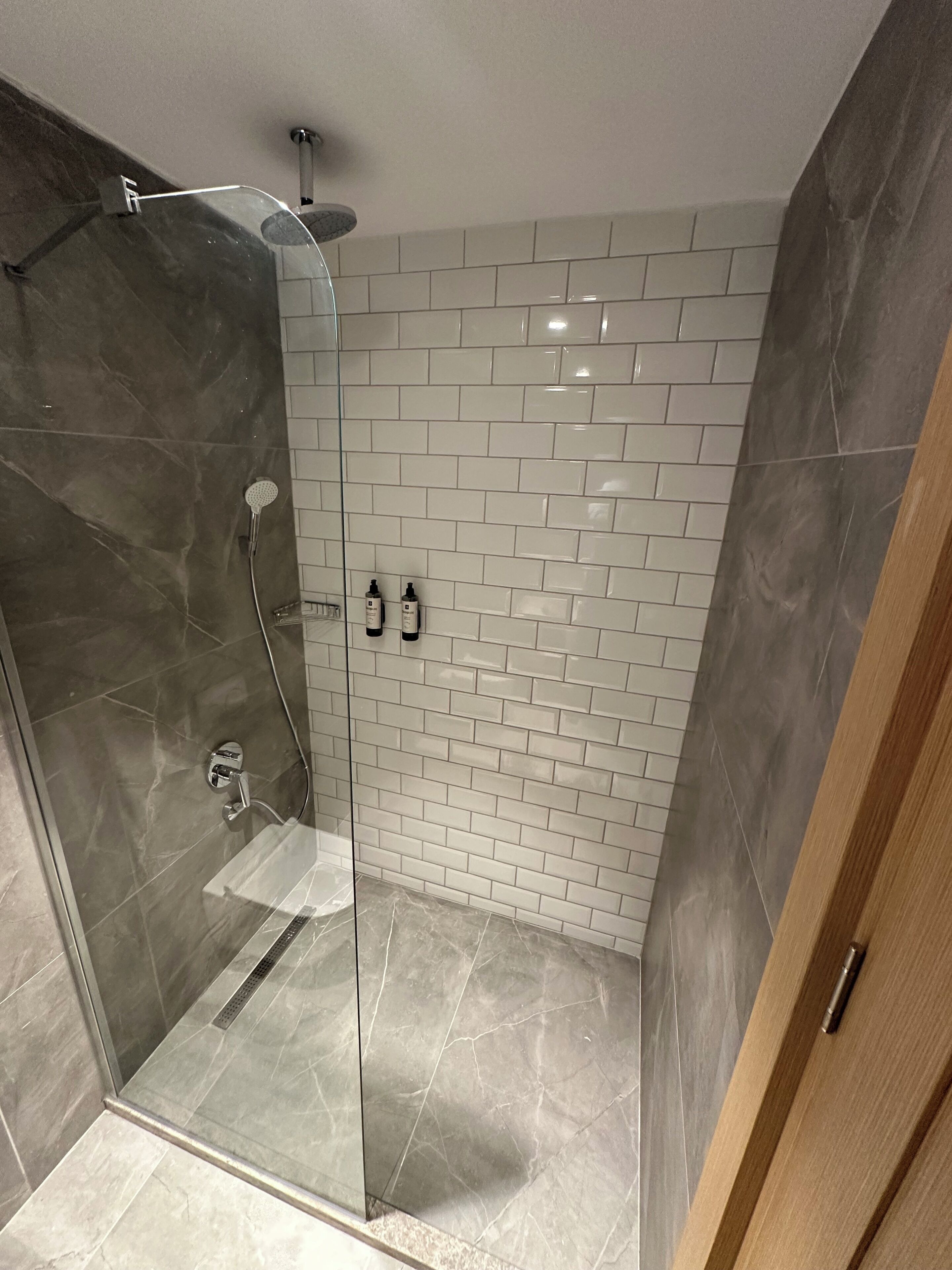 A glass-enclosed shower with a marble floor, white tiled walls, and a wooden door.
