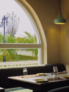 A restaurant with a large arched window, a green upholstered booth, and a table set for two. - Vijay Villas Rathara - Silpara Bypass Rd (Huzur)