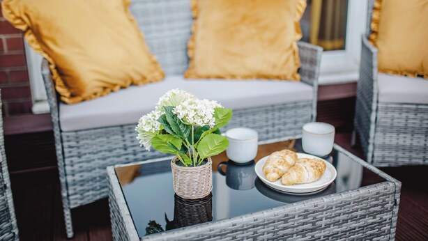 Móveis de vime com almofadas, uma mesa de vidro com uma planta, duas chávenas e um prato com pastelaria.