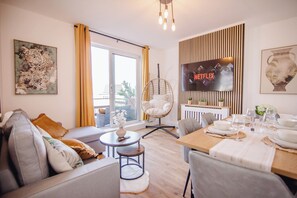 Ein modernes Wohnzimmer mit einer Couch, einem Esstisch für vier Personen, einem an der Wand montierten Fernseher und einem großen Fenster mit Vorhängen.