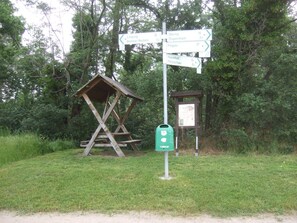 Ein Wegweiser mit mehreren Zielschildern, eine hölzerne Konstruktion, die einer Schutzhütte ähnelt, und eine grüne Infokasten.